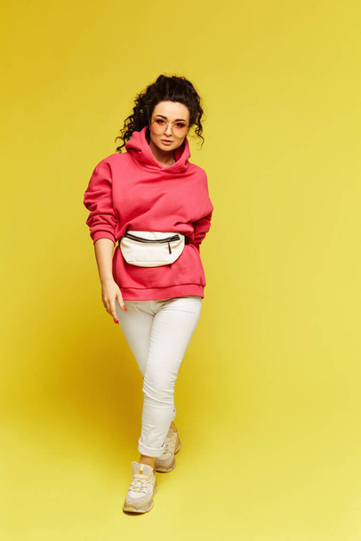 Model girl in pink sweatshirt and with belt bag walking over yellow background - Valokuva, kuva