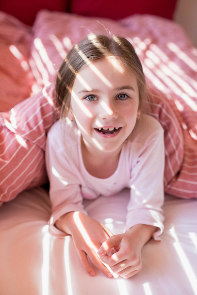 Cute little girl in bed woke up in the morning and smiles cheerfully. The sun's rays fall on the child's face through the blinds. Pastime at home during quarantine. Carefree childhood. Home comfort. - Foto, afbeelding