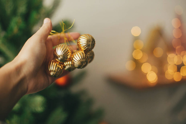 Hanging ornaments on christmas tree, festive decoration of room. Hand holding modern gold baubles on background of green tree. Decorating christmas tree. - Zdjęcie, obraz
