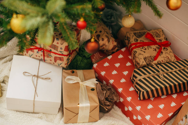 Καλά Χριστούγεννα. Κομψά χριστουγεννιάτικα δώρα κάτω από το χριστουγεννιάτικο δέντρο. Κουτιά δώρου τυλιγμένα σε μοντέρνο χαρτί και διακοσμημένο δέντρο με κόκκινα και χρυσά μπιχλιμπίδια στην αίθουσα εορταστικών σκανδάλων. - Φωτογραφία, εικόνα