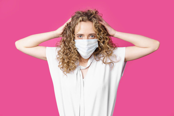 A young woman with a medical mask on her face holds her head with her hands. Isolated pink background - Foto, Imagen