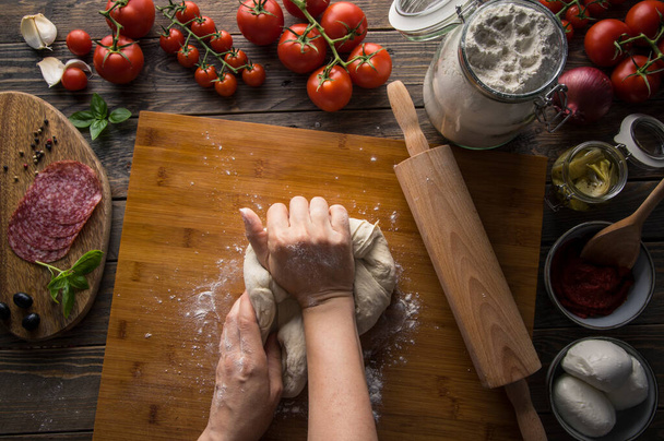 Una mujer que prepara una pizza, amasa la masa y pone ingredientes en la mesa de madera de la cocina. Oscuro y malhumorado, plano aéreo. - Foto, imagen