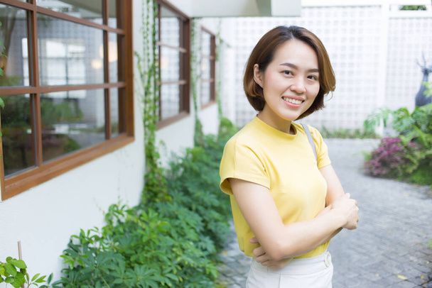 Femme asiatique aux cheveux courts dorés porte un t-shirt à manches courtes de couleur jaune et un pantalon crème alors qu'elle se tient debout et bras croisés dans le fond du jardin. - Photo, image
