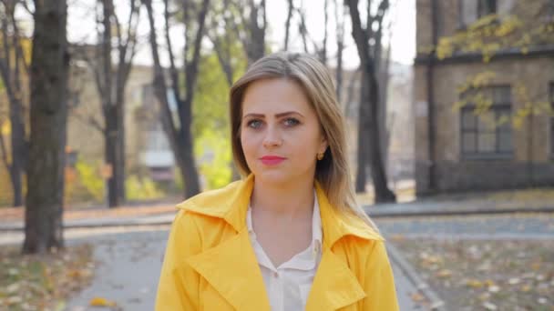 黄色のマントの女性は、白いブラウス笑みを浮かべて、秋の公園に立って、カメラを探して、カメラの女の子移動します。唇の上にピンクの口紅を持つ女の子屋外。4Kスローモーション映像 - 映像、動画