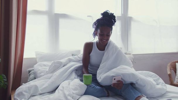 魅力的な若いアフリカ系アメリカ人の女性がベッドに座って、朝のコーヒーを飲み、電話を使用します - 写真・画像