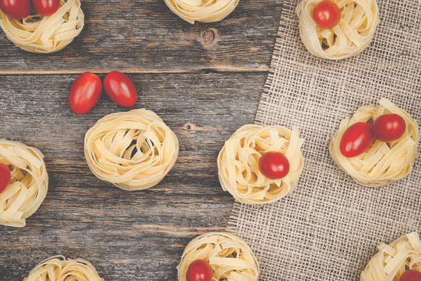 Nests of tagliatelle noodles with egg-shaped mini roma tomatoes on a rustic wooden table wit burlap, - Фото, изображение