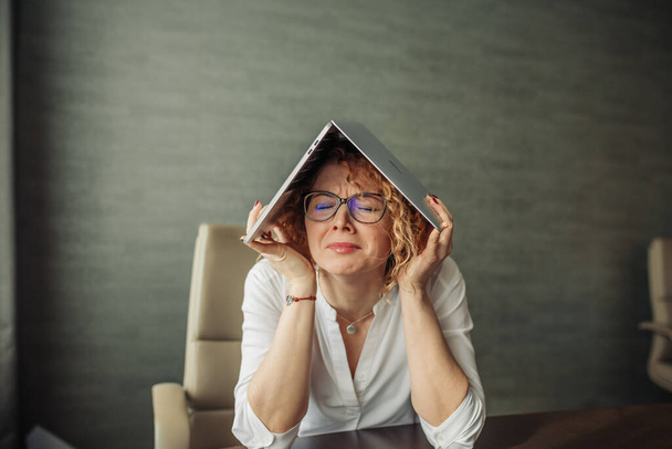 Tired woman with laptop on head at office with plaintive face and closed eyes - Foto, Imagen