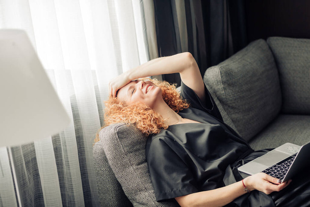 Red-haired woman with laptop having dreaming pleased face, relaxing on sofa. - Foto, afbeelding