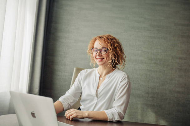 Happy red-haired woman in eyewear working on laptop sitting against grey wall - Photo, image