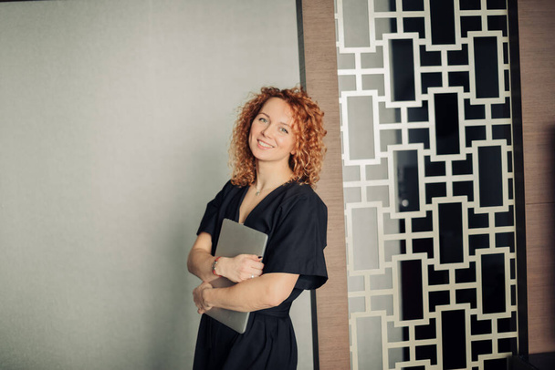 Woman in stylish black overalls with curly ginger hair posing with laptop - 写真・画像