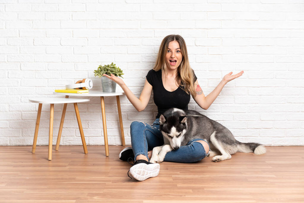 Jovem mulher bonita com seu cão husky sentado no chão em ambientes fechados com expressão facial chocada - Foto, Imagem
