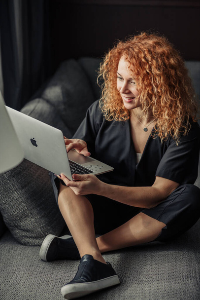 Happy redheaded woman entrepreneur works at laptop comfortably sitting at sofa. - Фото, изображение