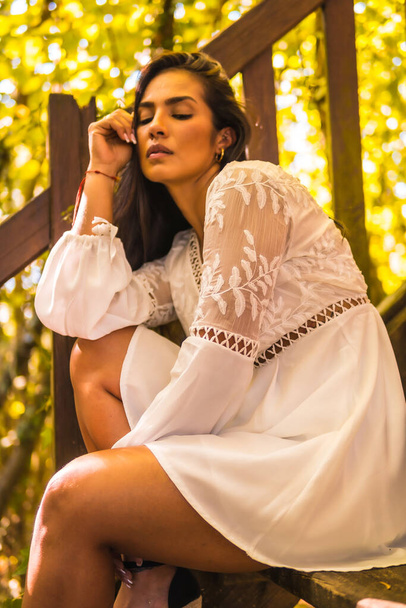 Lifestyle, brunette girl with straight hair in a beautiful white dress in a park resting in summer. Portrait sitting on a wooden ladder under trees - Photo, image