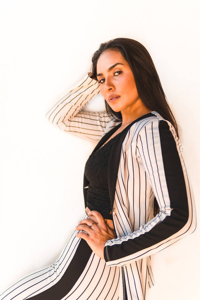Lifestyle, straight-haired Latin brunette girl in a white suit and black stripes. On a white background. Looking at the camera, in different postures, fashion editorial - Fotó, kép