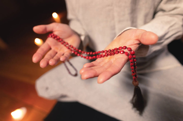 close-up man in clothes for practice and meditation sits in a lotus pose and holds red rosary to concentrate attention in a wooden room with dim light - Foto, Bild