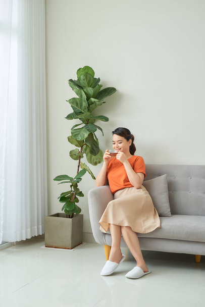 Portrait de jeune femme souriante avec tasse à thé assise sur le canapé à la maison - Photo, image