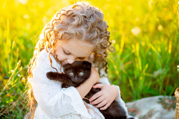klein mooi meisje knuffelt een zwart katje op het gazon in de zomer - Foto, afbeelding