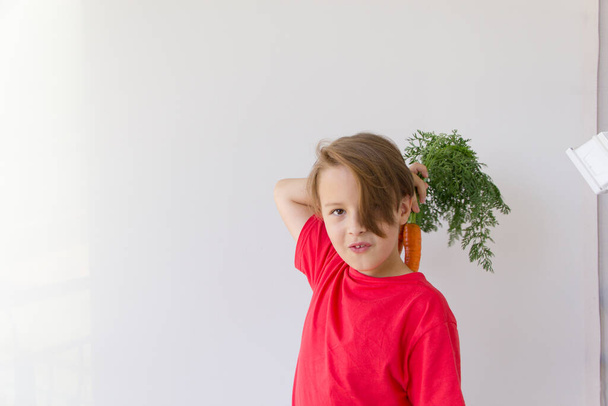 A cute boy in a red T-shirt holds a fresh carrot. Healthy food concept. - Zdjęcie, obraz