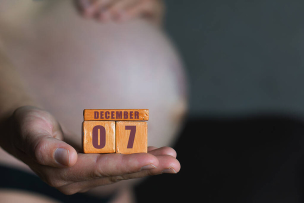 7 december. Dag 7 van de maand, geplande geboortedatum. Maand en dag geplaatst op houten kalender in zwangere vrouwen hand. wintermaand, dag van het jaar concept. - Foto, afbeelding