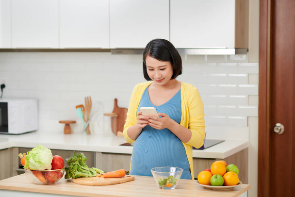 Беременная женщина на кухне готовит еду, используя мобильный телефон - Фото, изображение
