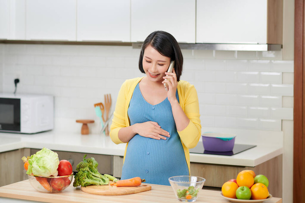Felice donna incinta chiamando su smartphone e facendo colazione in cucina a casa - Foto, immagini