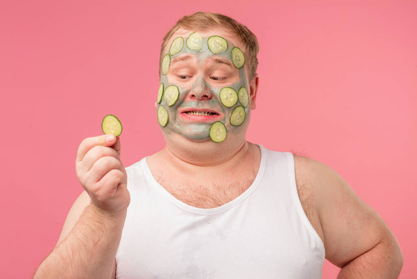Young man having doubts while applying cucumber mask on his face - Photo, image