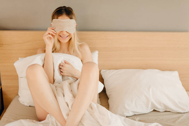 Ξανθιά γυναίκα κρύβει το σώμα της κάτω από κουβέρτα και τα μάτια της κάτω από μάσκα ύπνου - Φωτογραφία, εικόνα