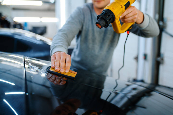 Mannelijke wikkel houdt squeegee en warmte pistool, auto tinting service. Werknemer die vinyl tint op het raam van het voertuig in de garage, getint auto-glas - Foto, afbeelding