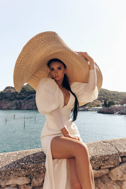 fashion  photo of beautiful woman with dark hair in luxurious white dress and hat posing in beautiful landscape with sea view - Photo, image