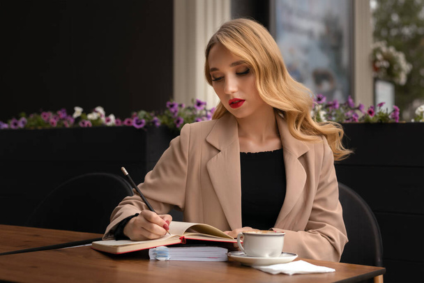 Молода красива жінка з довгим світлим волоссям п'є каву і пише щоденник, книгу або ноти у вуличному кафе, концепція способу життя
 - Фото, зображення