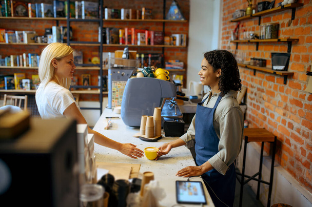 Женщина-бариста в фартуке дает кофе женщине в кафе. Девушка делает свежий эспрессо в кафетерии, официант за стойкой в баре - Фото, изображение