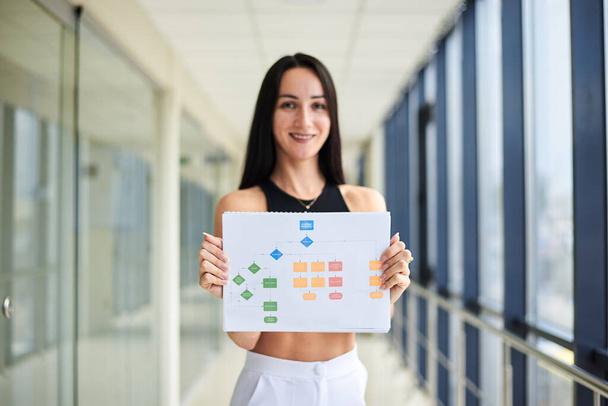 Young brunette woman, wearing casual business attire, holding white paper with diagram chart on it. Businesswoman, preparing for corporate presentation in light office building with huge windows. - Foto, Bild