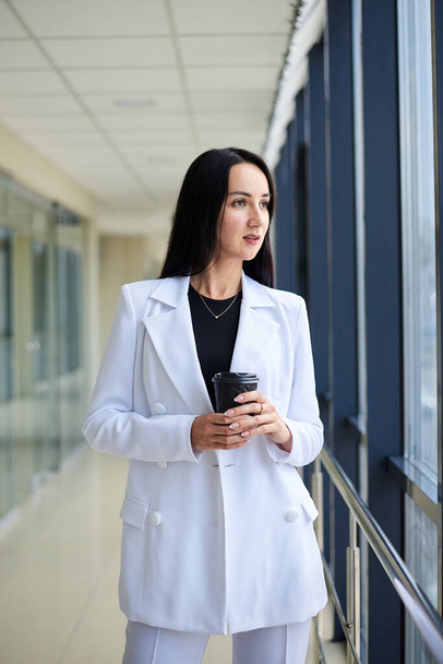 Young brunette woman, wearing white business suit, holding black paper coffee cup, standing in light office building near window. Businesswoman on lunch break. Corporate culture concept. - Φωτογραφία, εικόνα