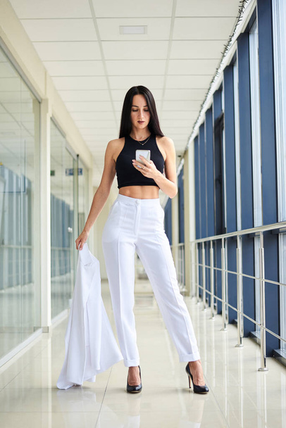 Young brunette woman, wearing white pants and black top, holding jacket in hand, taking selfie picture with cell phone in light passageway, posing for social media. Businesswoman on lunch break. - 写真・画像