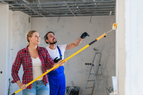 Mujer ayuda al hombre en overoles paredes de pintura con rodillo de pintura - Foto, Imagen