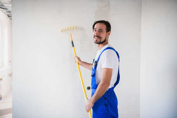 Craftsman paints the wall with a roller, smiles and looks at the camera - Fotoğraf, Görsel