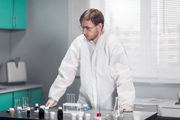 彼のテーブルにもたれて、研究室の真ん中に立っている化学保護服や眼鏡の男 - 写真・画像