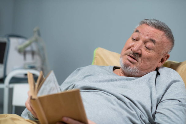 Un hombre mayor guapo leyendo un libro en la cama en el hospital - Foto, Imagen