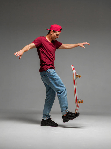 Cool guy skateboarder keeps skateboard on leg in studio on grey background. Photography about skateboarding and balance - Photo, image