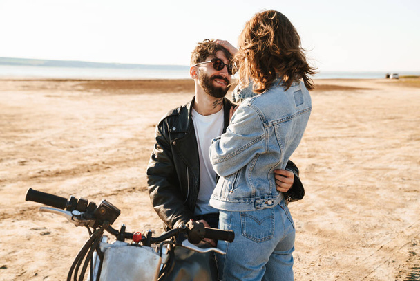 Attractive young happy couple embracing while leaning on a motorbike at the sunny beach - Foto, imagen
