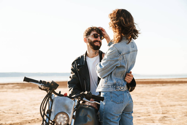 Attractive young happy couple embracing while leaning on a motorbike at the sunny beach - Φωτογραφία, εικόνα