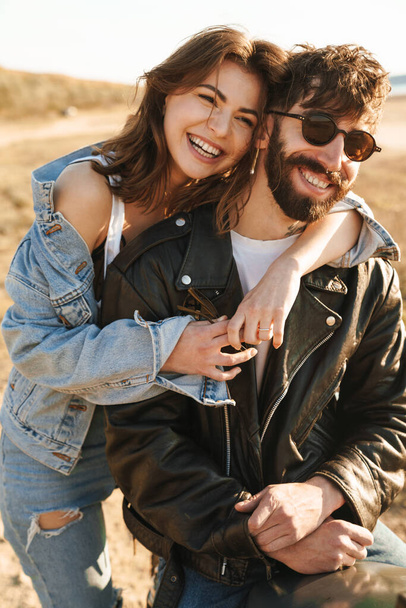 Attractive young happy couple embracing while leaning on a motorbike at the sunny beach - Photo, Image
