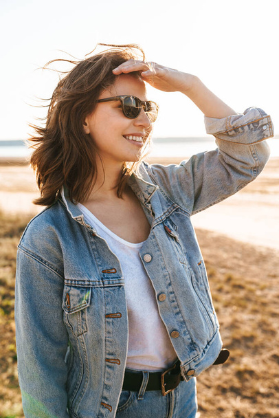 Красивая улыбающаяся молодая девушка в джинсовой куртке гуляет по солнечному пляжу - Фото, изображение