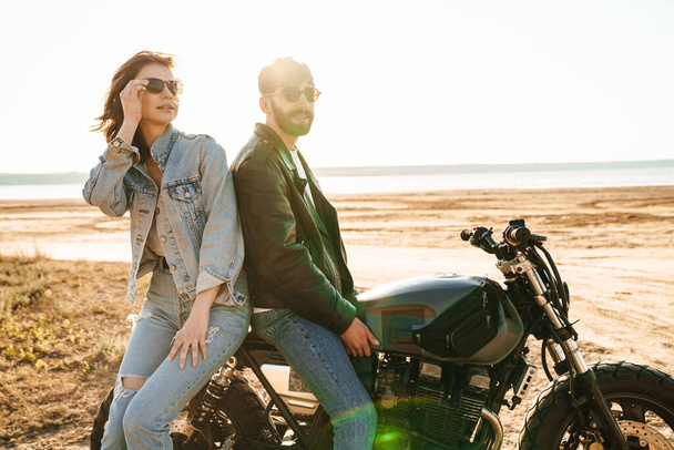 Attractive young happy couple embracing while leaning on a motorbike at the sunny beach - Photo, image