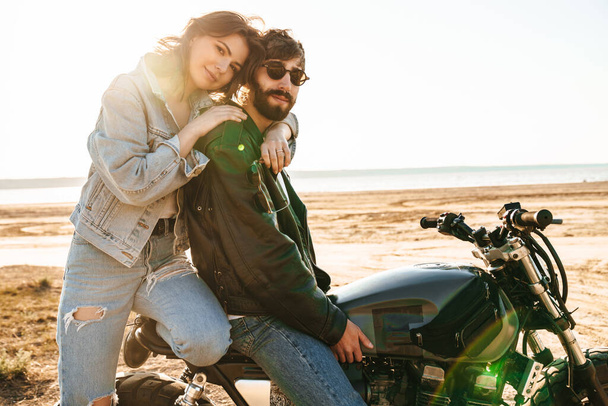 Beatiful yong couple wearing jackets sitting on a motorbike at the sunny beach, hugging - Photo, Image