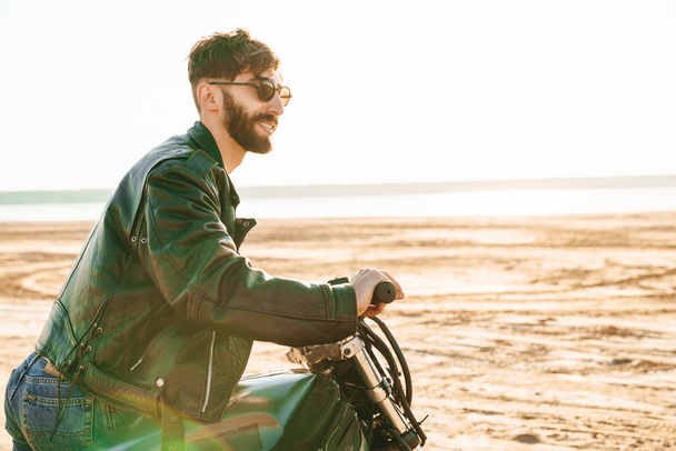 Handsome young bearded man wearing leather jacket sitting on a motorbike at the beach - Фото, изображение
