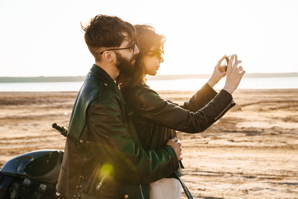 Attractive young happy couple embracing while leaning on a motorbike at the sunny beach, taking a selfie - Foto, immagini