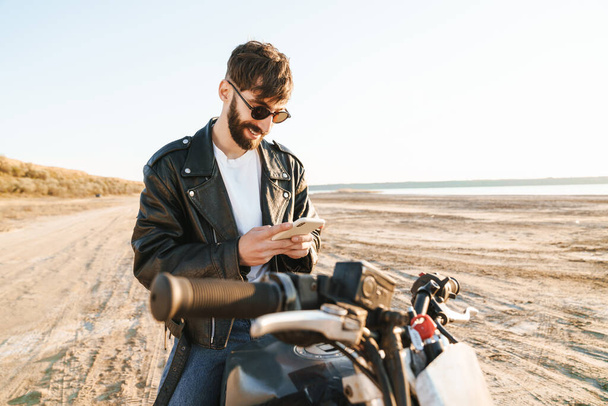 Handsome smiling young bearded man wearing leather jacket sitting on a motorbike at the beach, using mobile phone - Fotó, kép