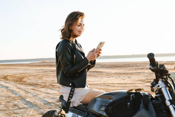 Beautiful young girl wearing leather jacket sitting on a motorbike at the sunny beach, using mobile phone - Фото, изображение