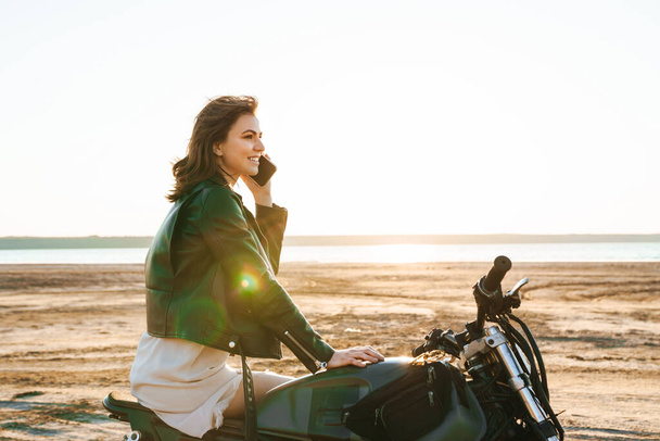 Beautiful young girl wearing leather jacket sitting on a motorbike at the sunny beach, talking on mobile phone - Zdjęcie, obraz